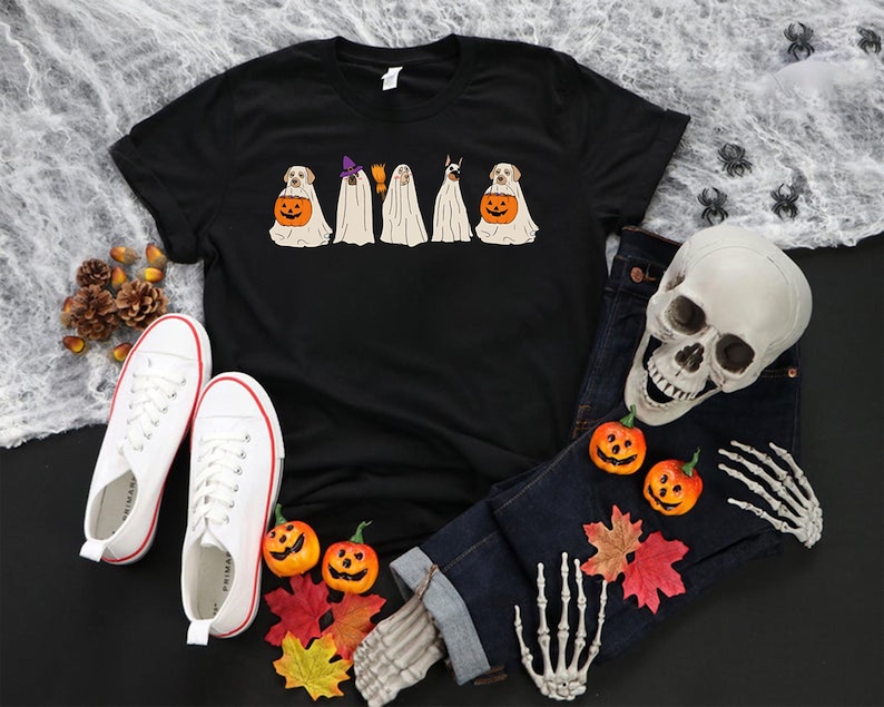 May include: Black t-shirt featuring a Halloween design with dogs dressed as ghosts, each holding a pumpkin. The shirt is paired with white sneakers, a pair of jeans, a skull, and decorative pumpkins and leaves.