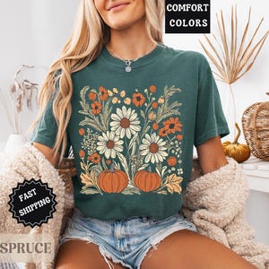Może przedstawiać: Turkusowa koszulka z kwiatowym wzorem z białymi stokrotkami, pomarańczowymi kwiatami i dwiema dyniami. Na koszulce widnieje napis "COMFORT COLORS". Widoczne są r&oacute;wnież słowa "BLUE SPRUCE" i "FAST SHIPPING".
