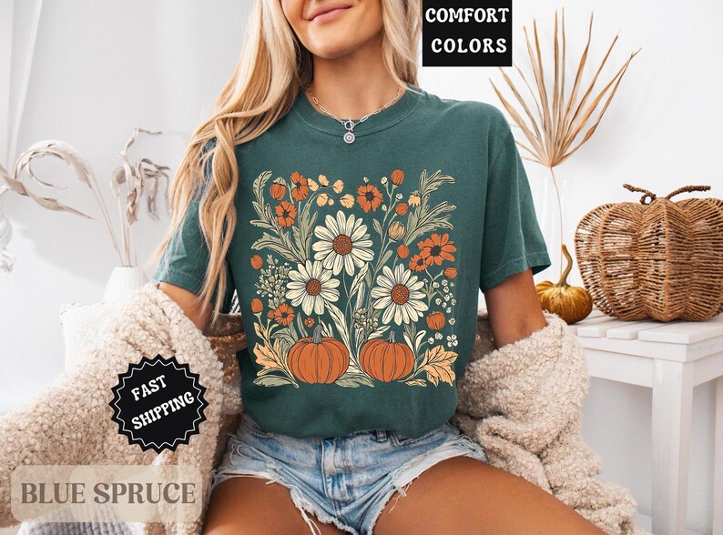 Może przedstawiać: Turkusowa koszulka z kwiatowym wzorem z białymi stokrotkami, pomarańczowymi kwiatami i dwiema dyniami. Na koszulce widnieje napis "COMFORT COLORS". Widoczne są r&oacute;wnież słowa "BLUE SPRUCE" i "FAST SHIPPING".