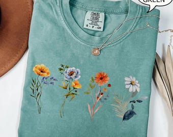 May include: A light green short-sleeve t-shirt featuring a graphic print of four distinct wildflower bouquets in yellow, blue, orange, and white. The shirt has a crew neckline and a Comfort Colors brand label at the inner collar. A thin silver chain necklace rests on the fabric.