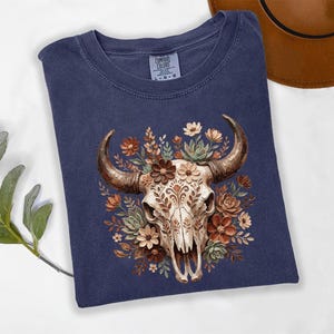 Puede incluir: Camiseta azul marino con una ilustraci&oacute;n detallada de una calavera de toro adornada con flores marrones, crema y verdes. La calavera tiene patrones intrincados. La camiseta est&aacute; hecha de un material suave y c&oacute;modo.