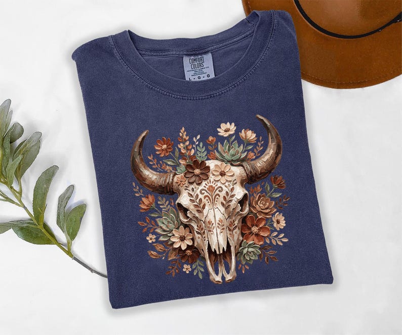 Puede incluir: Camiseta azul marino con una ilustraci&oacute;n detallada de una calavera de toro adornada con flores marrones, crema y verdes. La calavera tiene patrones intrincados. La camiseta est&aacute; hecha de un material suave y c&oacute;modo.