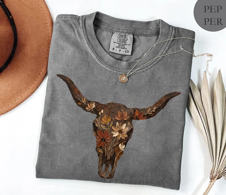 May include: A charcoal grey short-sleeve t-shirt featuring a graphic of a brown longhorn skull adorned with orange, cream, and brown flowers. The shirt has a crew neckline and a Comfort Colors brand label printed on the inside of the collar.