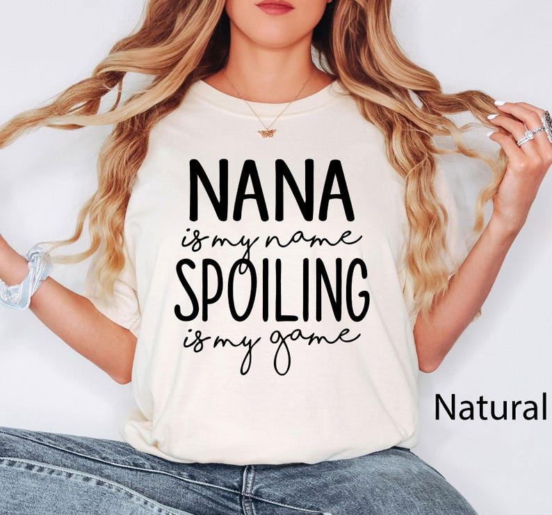 May include: Cream-colored t-shirt with the text "NANA is my name, SPOILING is my game" in black script. The word "Natural" is printed in a smaller font on the bottom right. The shirt is a casual style.