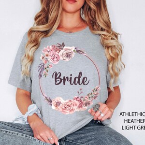Puede incluir: Camiseta gris claro con un dise&ntilde;o de corona floral y la palabra "Bride" en fuente cursiva. La corona presenta rosas rosas y acentos morados. La camiseta est&aacute; hecha de una tela suave y jaspeada.