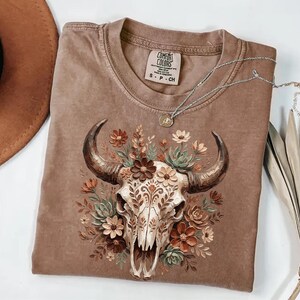 Puede incluir: Una camiseta Comfort Colors marr&oacute;n con un dise&ntilde;o de calavera de toro adornada con flores y hojas. La camiseta tiene cuello redondo y mangas cortas. Un collar plateado con un colgante est&aacute; drapeado sobre la camiseta.
