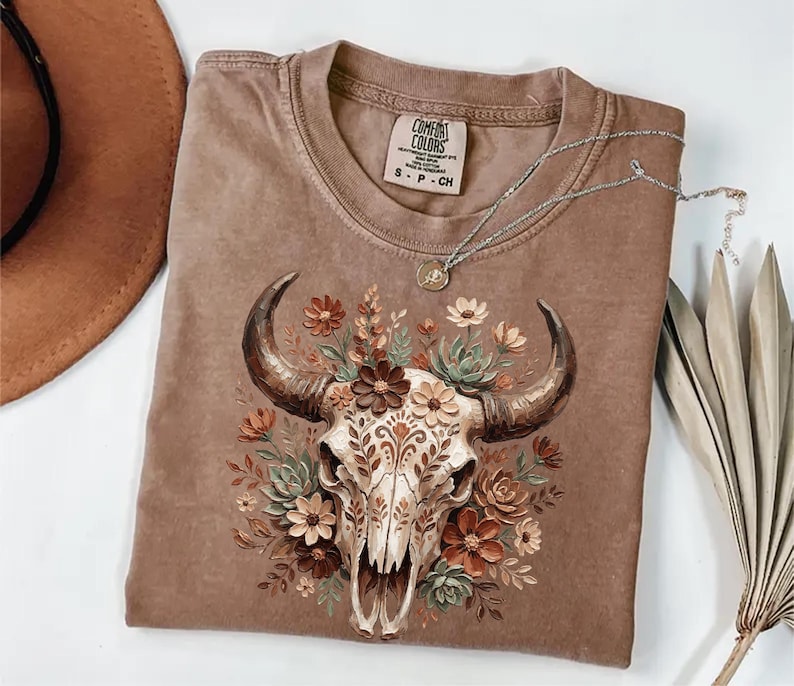 Puede incluir: Una camiseta Comfort Colors marr&oacute;n con un dise&ntilde;o de calavera de toro adornada con flores y hojas. La camiseta tiene cuello redondo y mangas cortas. Un collar plateado con un colgante est&aacute; drapeado sobre la camiseta.