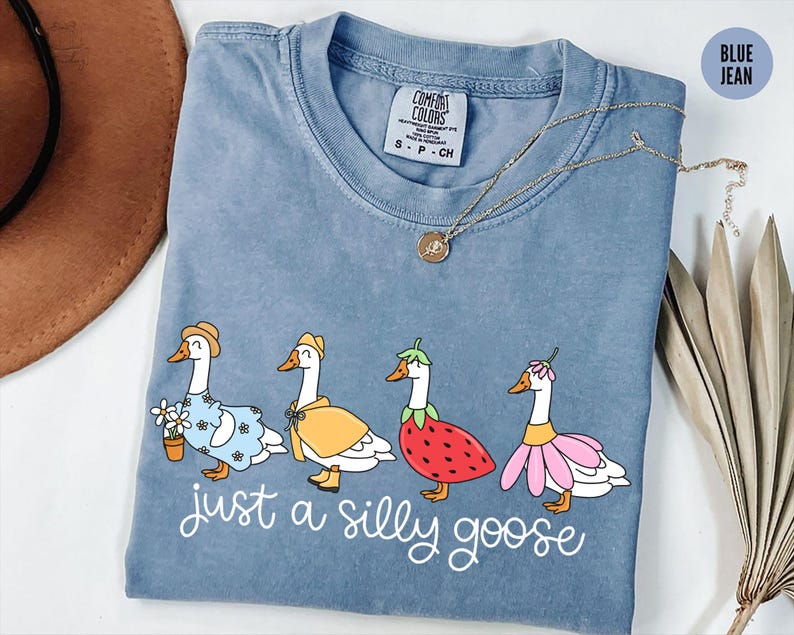 Puede incluir: Camiseta azul vaquero Comfort Colors con una ilustraci&oacute;n caprichosa de cuatro gansos vestidos con varios atuendos. El texto "just a silly goose" est&aacute; impreso debajo. Un collar dorado est&aacute; sobre la camiseta.