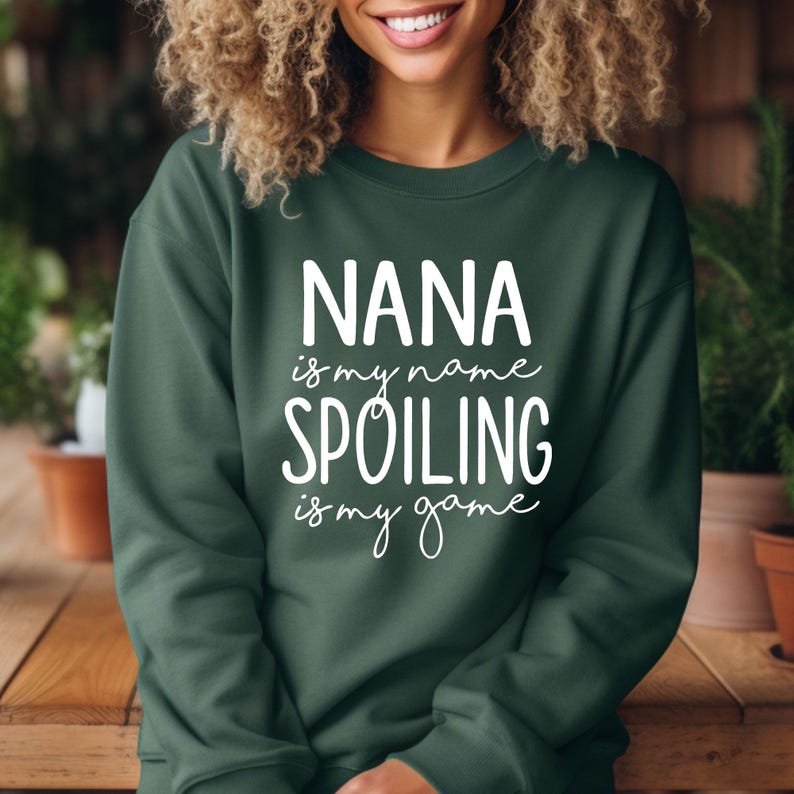 May include: Dark green sweatshirt with the text "NANA is my name SPOILING is my game" in white script. The sweatshirt is a crew neck style and appears to be made of a soft material. The text is centered on the front.