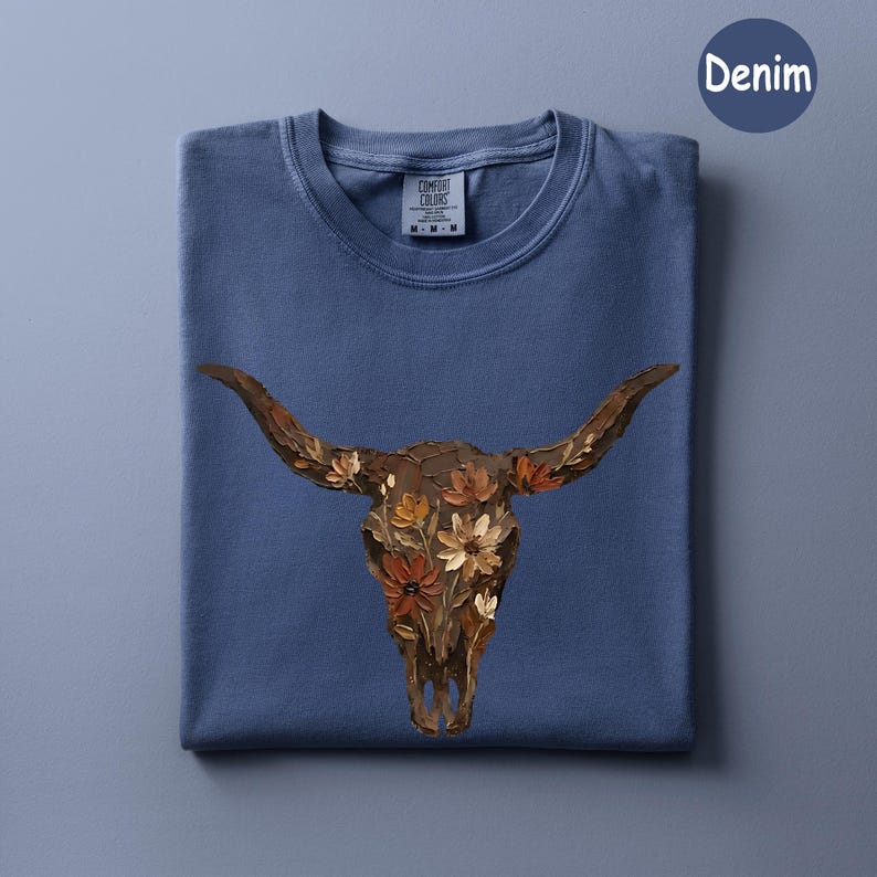 May include: A denim blue short-sleeve t-shirt featuring a graphic of a brown bull skull adorned with various orange, yellow, and brown flowers. The shirt has a crew neckline and a Comfort Colors brand label printed on the inside of the collar.