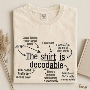 May include: An ivory t-shirt featuring black text that reads The shirt is decodable, surrounded by arrows and labels explaining linguistic concepts like Digraphs, Closed Syllable, r-controlled, and Latin-based prefixes and suffixes.