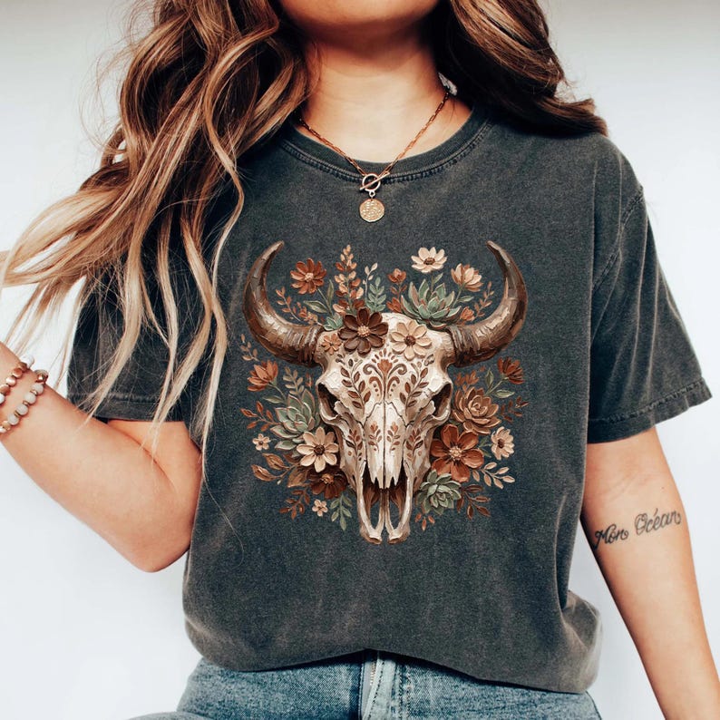 Puede incluir: Camiseta gris oscuro con una ilustraci&oacute;n detallada de una calavera de toro adornada con flores y follaje en tonos marr&oacute;n, beige y verde. El dise&ntilde;o est&aacute; centrado en la camiseta, creando una est&eacute;tica r&uacute;stica y bohemia.