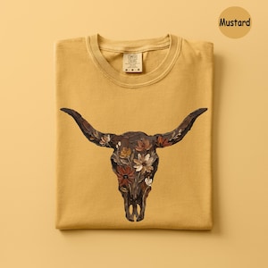 May include: A mustard yellow t-shirt featuring a graphic of a dark brown bull skull adorned with painted flowers in shades of white, cream, and rust orange. The shirt has a crew neckline and a Comfort Colors brand label printed on the inner collar.