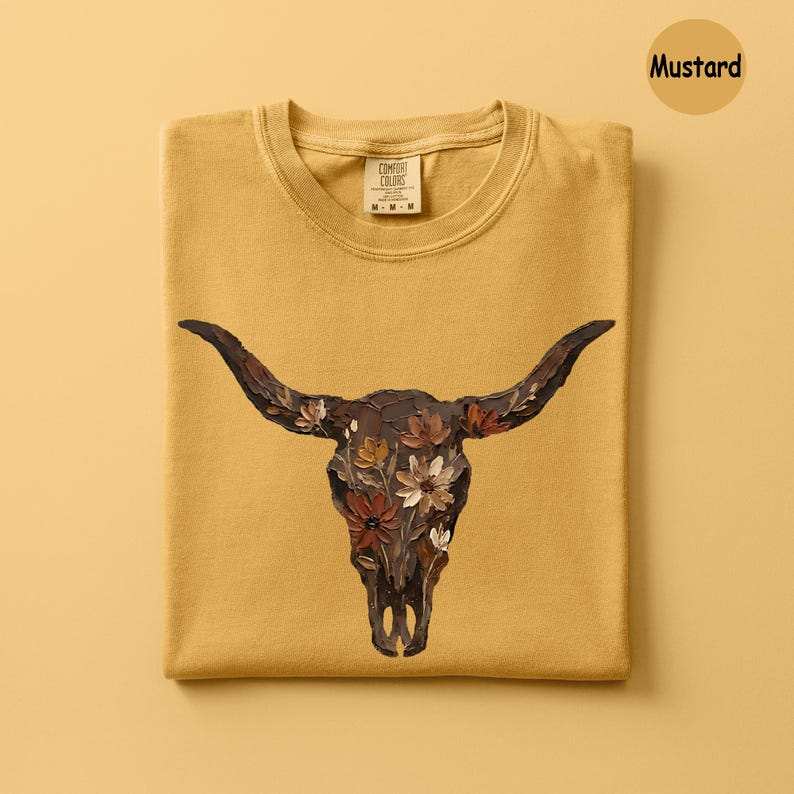 May include: A mustard yellow t-shirt featuring a graphic of a dark brown bull skull adorned with painted flowers in shades of white, cream, and rust orange. The shirt has a crew neckline and a Comfort Colors brand label printed on the inner collar.