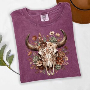 Puede incluir: Una camiseta de color ciruela con una ilustraci&oacute;n detallada de una calavera de toro adornada con elementos florales. La calavera es de color crema con patrones intrincados, rodeada de flores y suculentas marrones, beiges y verdes. La camiseta tiene un cuello redondo cl&aacute;sico.