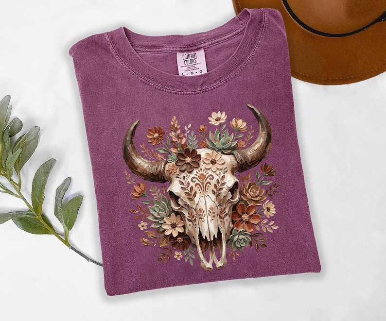 Puede incluir: Una camiseta de color ciruela con una ilustraci&oacute;n detallada de una calavera de toro adornada con elementos florales. La calavera es de color crema con patrones intrincados, rodeada de flores y suculentas marrones, beiges y verdes. La camiseta tiene un cuello redondo cl&aacute;sico.