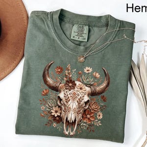 Puede incluir: Camiseta color c&aacute;&ntilde;amo con una ilustraci&oacute;n detallada de una calavera de toro adornada con flores en tonos marr&oacute;n, crema y naranja. La camiseta tiene cuello redondo cl&aacute;sico y mangas cortas. Un collar plateado con un colgante descansa sobre la camiseta.