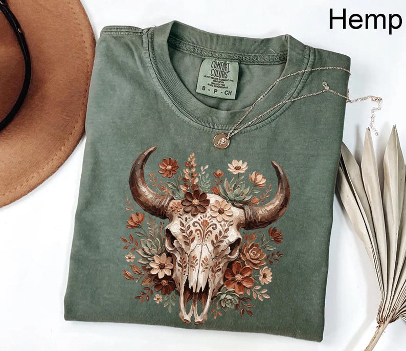 Puede incluir: Camiseta color c&aacute;&ntilde;amo con una ilustraci&oacute;n detallada de una calavera de toro adornada con flores en tonos marr&oacute;n, crema y naranja. La camiseta tiene cuello redondo cl&aacute;sico y mangas cortas. Un collar plateado con un colgante descansa sobre la camiseta.