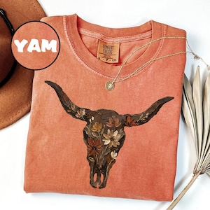 May include: A terracotta-coloured t-shirt featuring a graphic of a dark brown cow skull adorned with painted flowers. The shirt has a crew neckline and a Comfort Colors brand label at the inner neck. A circular inset in the top left corner displays the text YAM.
