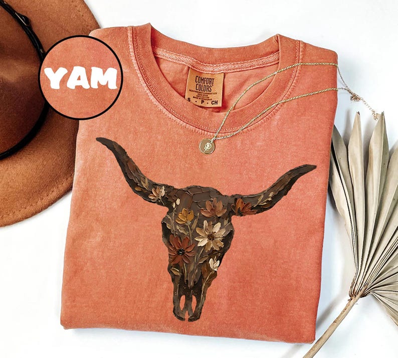May include: A terracotta-coloured t-shirt featuring a graphic of a dark brown cow skull adorned with painted flowers. The shirt has a crew neckline and a Comfort Colors brand label at the inner neck. A circular inset in the top left corner displays the text YAM.