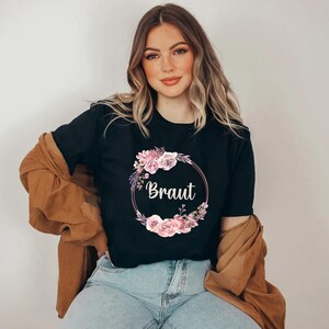 Puede incluir: Camiseta negra con la palabra "Braut" en escritura blanca, rodeada por una corona floral de flores rosas y moradas. La modelo lleva vaqueros azules y una chaqueta marr&oacute;n.