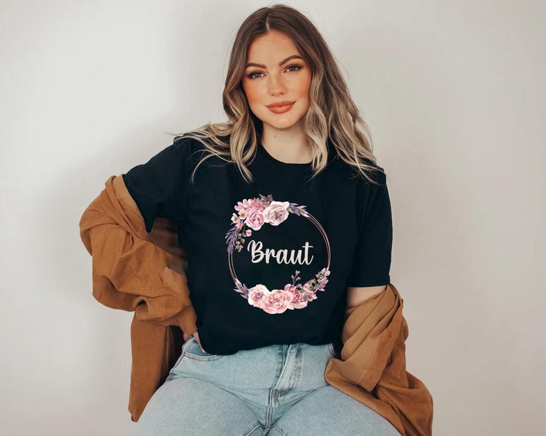 Puede incluir: Camiseta negra con la palabra "Braut" en escritura blanca, rodeada por una corona floral de flores rosas y moradas. La modelo lleva vaqueros azules y una chaqueta marr&oacute;n.