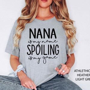 May include: Light grey t-shirt with the text "NANA is my name, SPOILING is my game" in black. The shirt is a heathered fabric and has short sleeves. The text "ATHLETHICH HEATHER LIGHT GREY" is printed on the right side of the shirt.