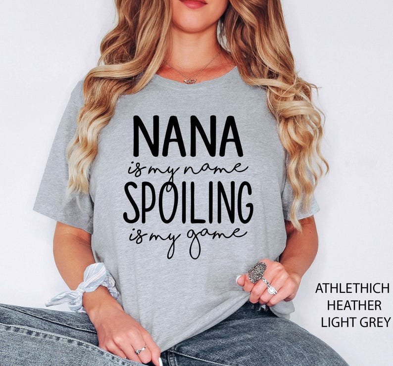 May include: Light grey t-shirt with the text "NANA is my name, SPOILING is my game" in black. The shirt is a heathered fabric and has short sleeves. The text "ATHLETHICH HEATHER LIGHT GREY" is printed on the right side of the shirt.
