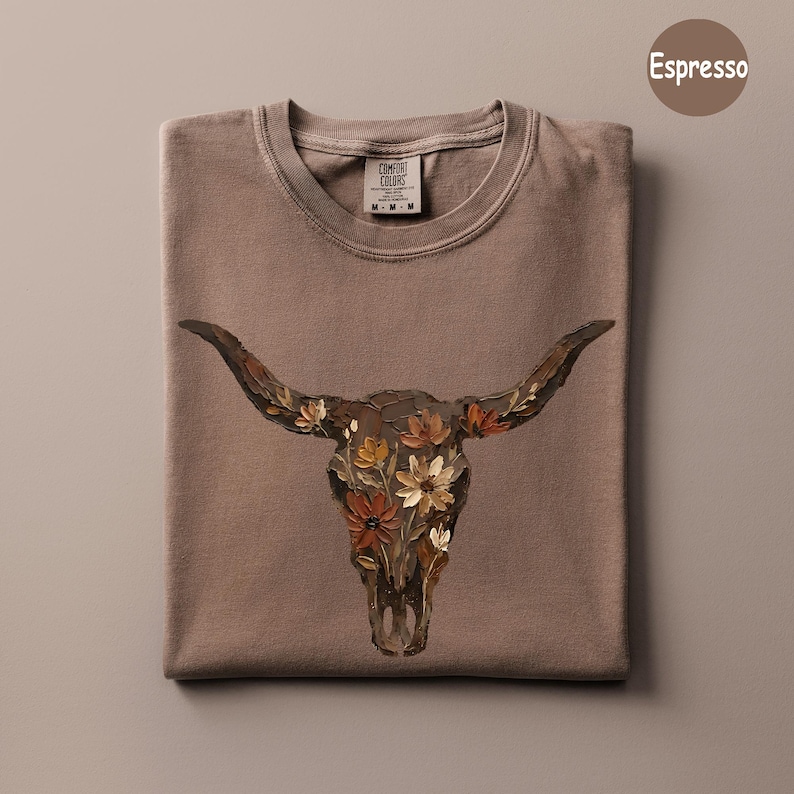 May include: An espresso-coloured t-shirt featuring a graphic of a longhorn skull adorned with painted flowers in shades of orange, yellow, and white. The shirt has a crew neckline and a Comfort Colors brand label at the inner neck area.