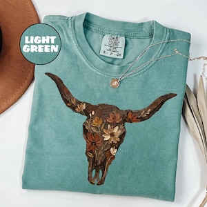 May include: A light green short-sleeved t-shirt featuring a graphic of a brown cow skull adorned with orange, yellow, and brown flowers. The shirt has a crew neckline and a Comfort Colors brand label on the inside collar with the text S P CH.