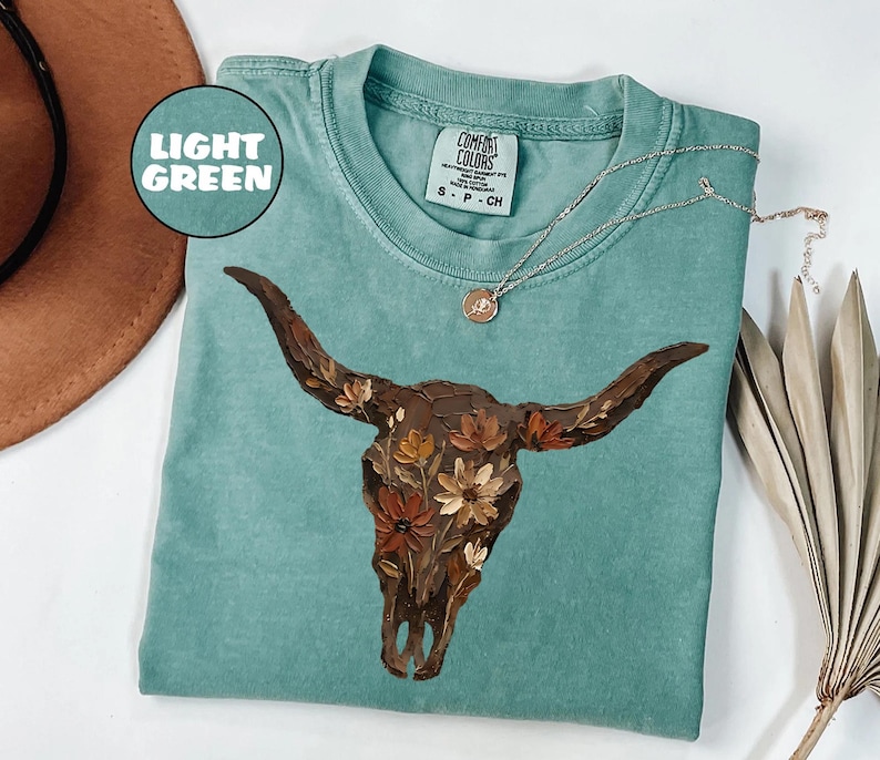 May include: A light green short-sleeved t-shirt featuring a graphic of a brown cow skull adorned with orange, yellow, and brown flowers. The shirt has a crew neckline and a Comfort Colors brand label on the inside collar with the text S P CH.