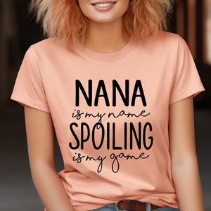May include: A peach-colored t-shirt with the text "NANA is my name, SPOILING is my game" in black script. The shirt has a crew neck and short sleeves. The person wearing the shirt is smiling.