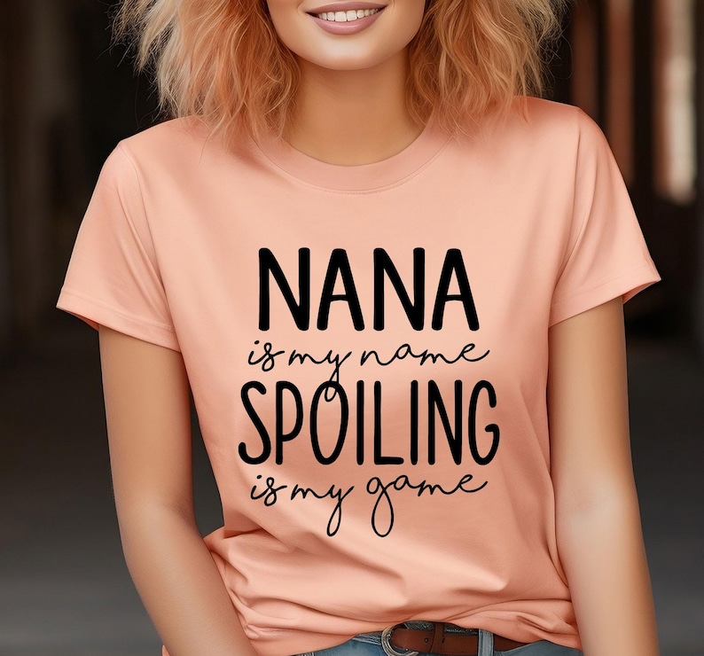 May include: A peach-colored t-shirt with the text "NANA is my name, SPOILING is my game" in black script. The shirt has a crew neck and short sleeves. The person wearing the shirt is smiling.