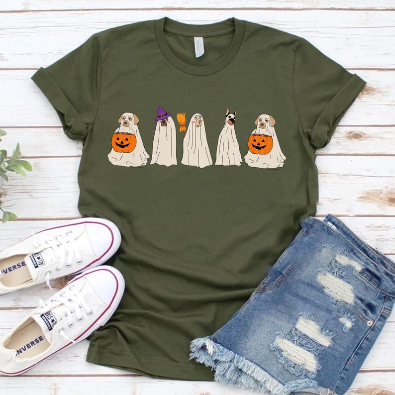 May include: Olive green t-shirt featuring a Halloween design with five dogs dressed as ghosts. Each ghost dog is holding a jack-o'-lantern. The shirt is paired with white sneakers and denim shorts.