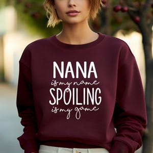May include: A maroon sweatshirt with the text "NANA is my name SPOILING is my game" in white script. The sweatshirt is a solid color and has a crew neck.