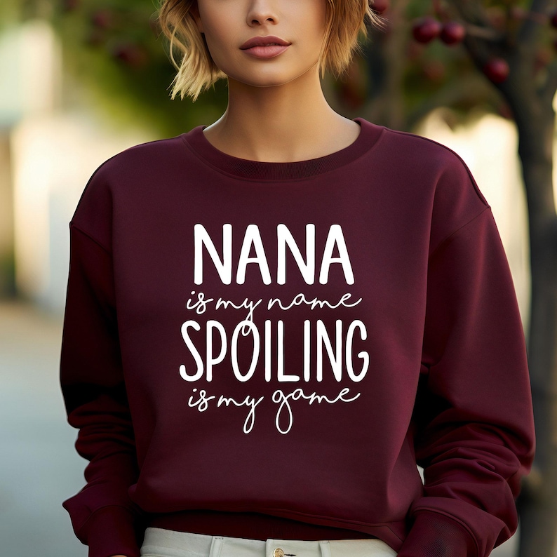 May include: A maroon sweatshirt with the text "NANA is my name SPOILING is my game" in white script. The sweatshirt is a solid color and has a crew neck.