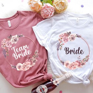 Puede incluir: Dos camisetas, una rosa empolvado y otra blanca, con dise&ntilde;os de coronas florales. La camiseta rosa empolvado dice "Team Bride" y la camiseta blanca dice "Bride". Ambas camisetas tienen un dise&ntilde;o de corona floral redonda con rosas rosas y melocot&oacute;n.