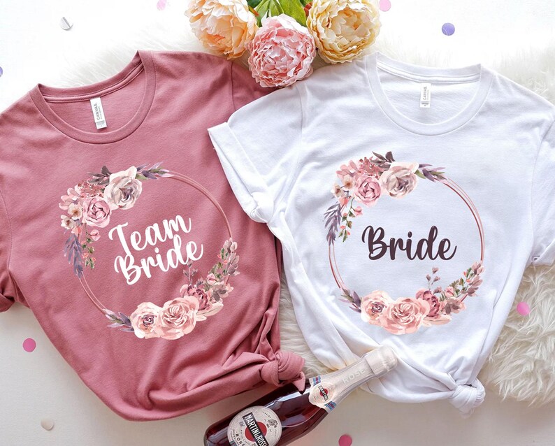Puede incluir: Dos camisetas, una rosa empolvado y otra blanca, con dise&ntilde;os de coronas florales. La camiseta rosa empolvado dice "Team Bride" y la camiseta blanca dice "Bride". Ambas camisetas tienen un dise&ntilde;o de corona floral redonda con rosas rosas y melocot&oacute;n.