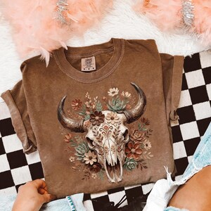 Puede incluir: Camiseta marr&oacute;n con un dise&ntilde;o de calavera de toro adornada con flores y hojas. La calavera tiene patrones detallados, y las flores son en tonos marr&oacute;n, crema y verde. La camiseta tiene un corte relajado y cuello redondo.
