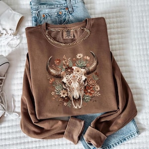 Puede incluir: Una sudadera marr&oacute;n con un dise&ntilde;o de calavera de toro floral y un collar de cadena dorada. La imagen muestra la sudadera junto con unos vaqueros azules y zapatillas blancas, ofreciendo un estilo relajado y bohemio.