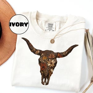 May include: An ivory short-sleeve t-shirt featuring a graphic of a dark brown bull skull adorned with various orange, yellow, and brown flowers. The shirt has a Comfort Colors brand label at the neck with the text Ivory in a circular graphic.