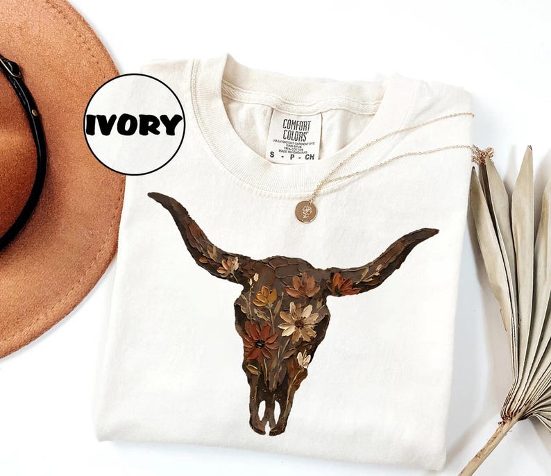 May include: An ivory short-sleeve t-shirt featuring a graphic of a dark brown bull skull adorned with various orange, yellow, and brown flowers. The shirt has a Comfort Colors brand label at the neck with the text Ivory in a circular graphic.