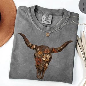 May include: A charcoal grey short-sleeve t-shirt featuring a graphic of a brown longhorn skull adorned with orange, cream, and brown flowers. The shirt has a crew neckline and a Comfort Colors brand label printed on the inside of the collar.