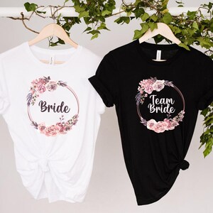 Puede incluir: Dos camisetas, una blanca y otra negra, con dise&ntilde;os de coronas florales. La camiseta blanca dice "Bride", la negra "Team Bride". Ambas cuelgan de perchas de madera, con follaje en el fondo.