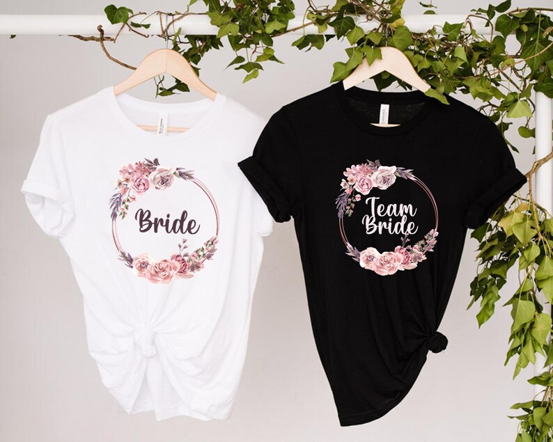 Puede incluir: Dos camisetas, una blanca y otra negra, con dise&ntilde;os de coronas florales. La camiseta blanca dice "Bride", la negra "Team Bride". Ambas cuelgan de perchas de madera, con follaje en el fondo.