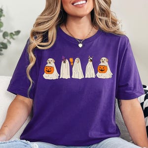 May include: A purple t-shirt featuring a Halloween-themed graphic. The design shows five dogs dressed as ghosts. Two dogs are holding orange jack-o'-lanterns. One ghost dog is wearing a witch hat and holding a broom.