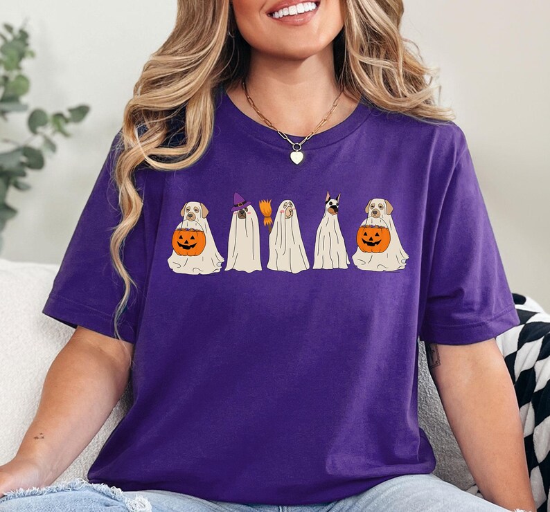 May include: A purple t-shirt featuring a Halloween-themed graphic. The design shows five dogs dressed as ghosts. Two dogs are holding orange jack-o'-lanterns. One ghost dog is wearing a witch hat and holding a broom.