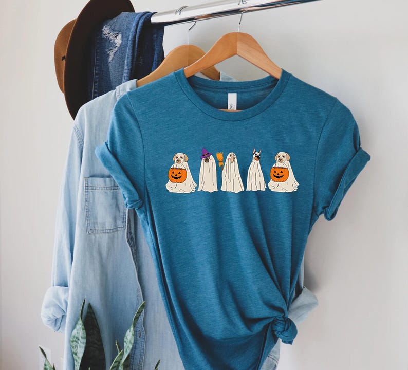 May include: Teal blue t-shirt featuring a Halloween-themed design. The graphic depicts several dogs dressed as ghosts, each with a unique hat or holding a jack-o'-lantern. The shirt is displayed on a wooden hanger with other clothing items.