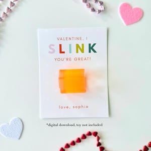 May include: A Valentine's Day card with the text "VALENTINE, I SLINK YOU'RE GREAT!" and "love, sophie". An orange toy is on the card. Pink, red, and white heart-shaped decorations surround the card. The text "*digital download, toy not included" is also present.