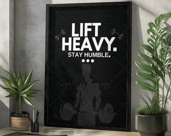 Puede incluir: Póster enmarcado en negro con el texto "LIFT HEAVY. STAY HUMBLE." en blanco. Una silueta de un levantador de pesas está en el centro. El póster está sobre un escritorio de madera con una planta en maceta a la izquierda y a la derecha.